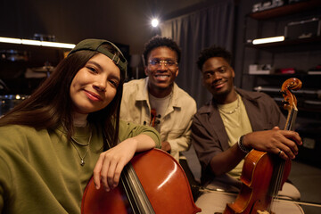 Portrait of young adult Hispanic woman holding cello taking selfie with two young adult Black men,...