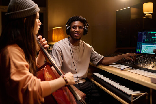 Black young adult man wearing headphones operating audio mixing console while Hispanic young adult woman playing cello in recording studio, both collaborating on music production project