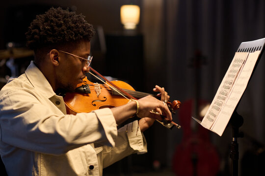 Young adult Black man playing violin while reading sheet music on stand, focusing intently on performance in indoor setting, demonstrating musical skill and concentration