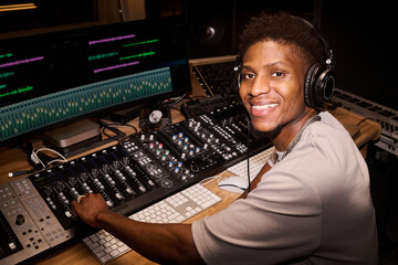 Portrait of young adult Black man wearing headphones smiling at camera, while operating...