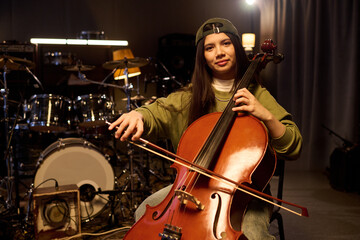 Portrait of Latin female musician sitting in music studio playing cello, holding bow and looking...