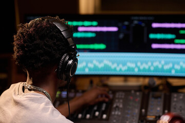 Black young adult man wearing headphones operating audio mixing console in music studio, adjusting...