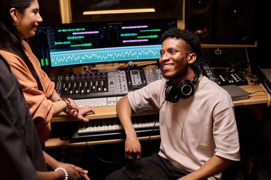 Young Black man sitting in music studio wearing headphones around neck, smiling and interacting with young Hispanic woman while surrounded by audio mixing equipment and computer monitors