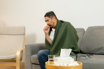 A young man with a fever lying under a blanket on the sofa, resting at home for recovery.