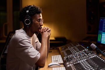 Young adult Black man wearing headphones sitting at mixing console working in professional...