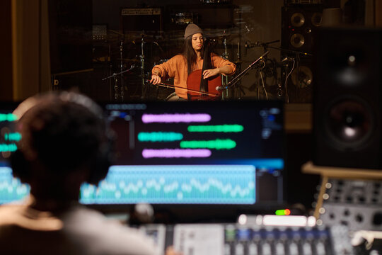Young adult Latin woman playing cello in professional recording studio, sitting behind microphone with music producer monitoring audio levels on mixing console in foreground - Powered by Adobe