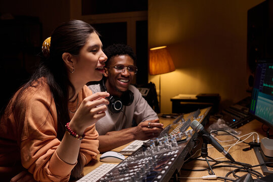 Young adult Hispanic woman and young adult Black man working together at audio mixing console in recording studio, both smiling and focusing on computer monitor during music production