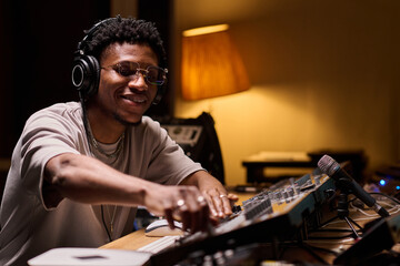 Young adult Black man wearing headphones operating audio mixing console in recording studio, smiling while adjusting controls, professional music production equipment visible on desk