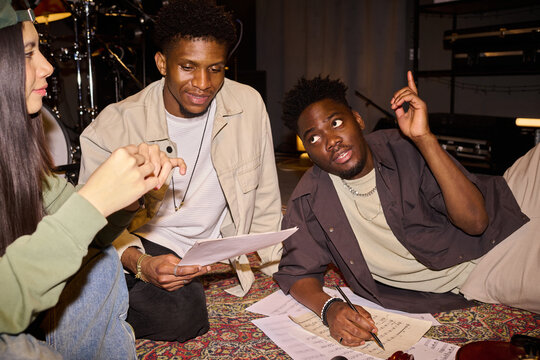 Young Black man lying on floor holding pen and gesturing while discussing music sheets with young African American man and young Hispanic woman sitting nearby, collaborating on creative project - Powered by Adobe