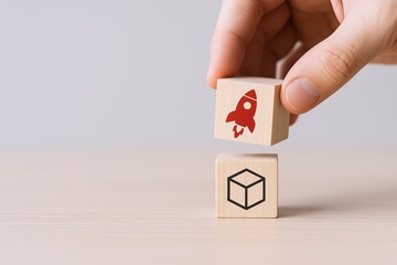 Hand stacking two wooden blocks with rocket and cube illustrations on a plain background.