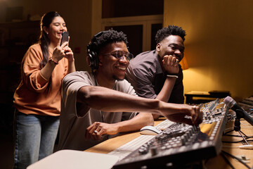Young Black man operating audio mixing console in recording studio, smiling while young Black man...