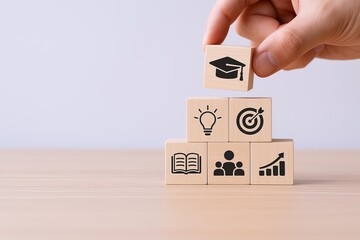 Hand holding wooden block with graduation cap symbol above stack of educational and growth icons arranged on light wood surface.