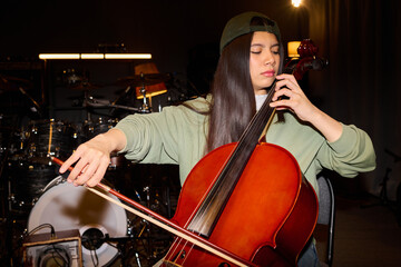 Hispanic girl playing cello in music studio, sitting with focused expression, long straight hair visible under cap, holding bow and instrument, drums in background