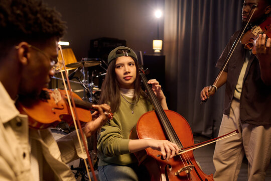 Latin girl playing cello surrounded by Black young men playing violins in music studio, focused expressions, musical collaboration, instruments visible in background