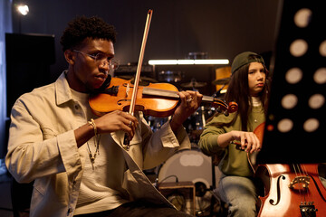 Young Black man playing violin alongside Hispanic girl playing cello in music studio, both focused...