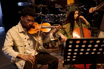 Black man playing violin and Hispanic girl playing cello in music studio, both focused on instruments, music stand with sheet music in foreground, drum set in background