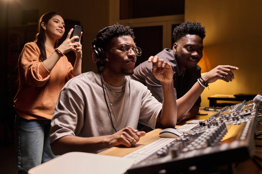 Two young adult Black men sitting at audio mixing console working on music production, while young adult Hispanic woman standing behind recording video on smartphone in recording studio