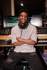 Portrait of young adult Black man sitting in music studio, smiling at camera wearing headphones...