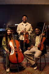Multiethnic group of young adult musicians posing with string instruments in music studio, Latin...