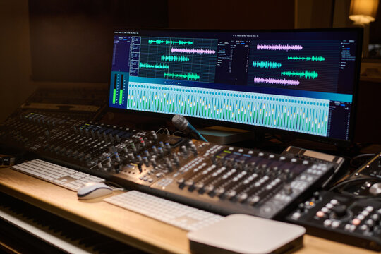 Professional audio mixing console with multiple sliders and buttons positioned in front of large computer monitor displaying digital audio workstation interface with sound waveforms - Powered by Adobe