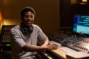 Young adult Black man sitting at audio mixing console working on music production in professional...