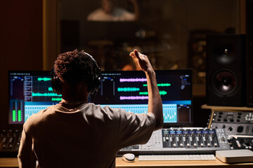 Black young adult man wearing headphones working at audio mixing console in professional recording...