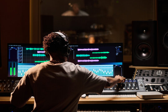 Black young adult man wearing headphones working at audio mixing console in professional recording studio, adjusting sound levels on computer monitors with music producer in background