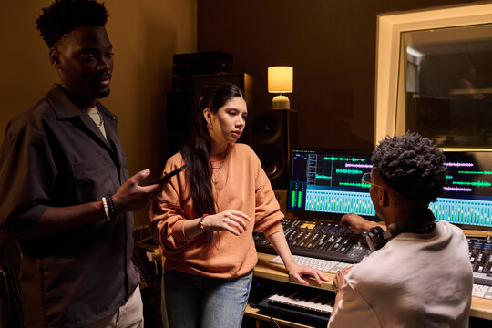 Multiethnic group of young adults collaborating in music studio, Black man holding smartphone, Hispanic woman and Black man working with professional audio mixing equipment, discussing project