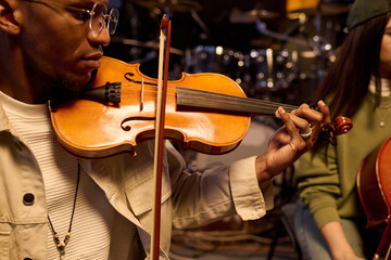 Black young adult man playing violin in music studio, focusing on instrument while sitting next to...