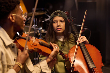 Hispanic girl playing cello while looking at young Black man playing violin in music studio, both...