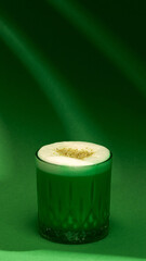 Pistachio latte close-up with thick foam and nut garnish on deep green background. Concept of beverage photography, cafe branding, winter menu, cozy stylish drinks.