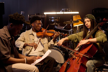 Black young adult man playing violin next to Latin young adult woman playing cello while sitting in...