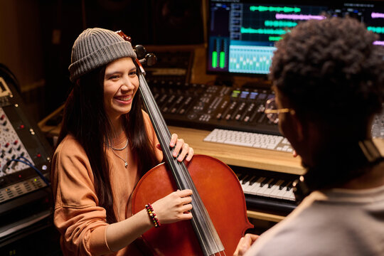 Young adult Hispanic woman smiling while playing cello in music studio, interacting with young adult Black man sitting nearby, professional audio equipment visible in background