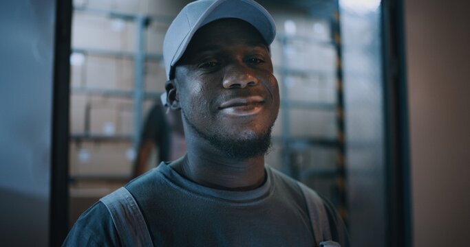Outside of Logistics Distribution Warehouse at Night: Portrait of African American Employee Looking at Camera, Working in Delivery Service. Loaders Carrying Packages in the Background. Close Up Shot.