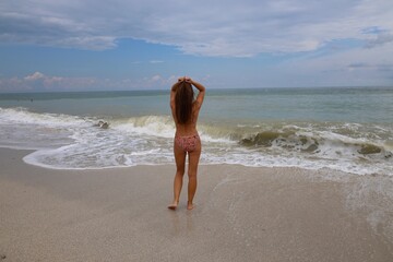 young woman stands on the beach with her back to the camera, wearing a bikini and gazing at the...