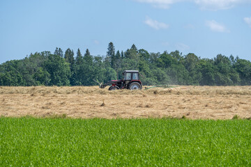Traditional farm tractor working on hayfield under blue sky, agricultural landscape showing summer...