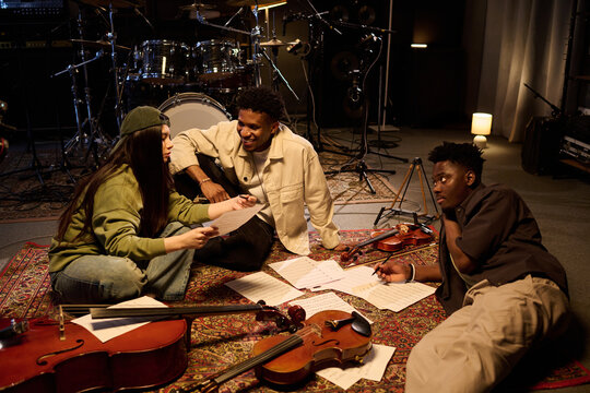Multiethnic group of young adults sitting on floor in music studio discussing sheet music surrounded by violins and cello, collaborating on musical arrangement, smiling and interacting