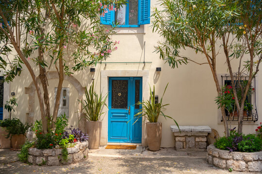 A charming traditional house in Osor, Croatia, featuring bright blue shutters and doors, surrounded by potted plants and oleander trees