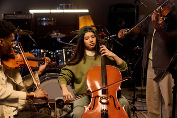 Latin girl playing cello with eyes closed surrounded by Black men playing violins in music studio,...