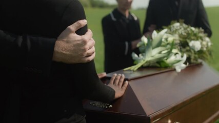 Cropped slowmo of hand supporting widow in mourning standing near wooden coffin with white lilies on it, grieving loss of loved one at outdoor funeral ceremony
