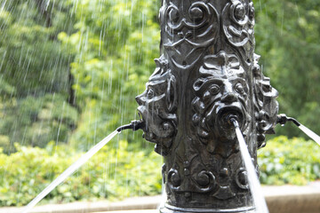 Rain falls around an ornate cast iron fountain with lion-like faces, each spouting water.