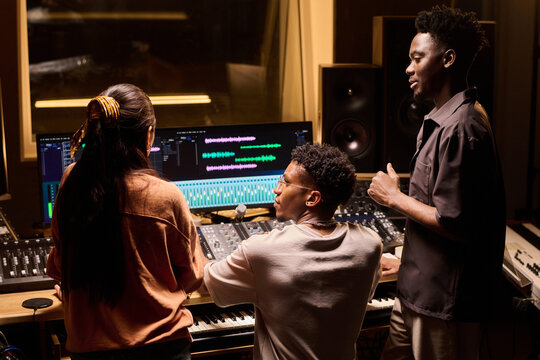 Young Black man operating audio mixing console while young Black man and young woman standing nearby discussing music production in professional recording studio