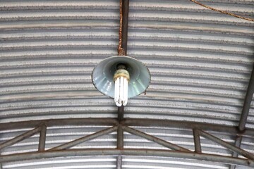 A ceiling-mounted lamp with a funnel-shaped shade hangs beneath a market roof. The roof is designed...