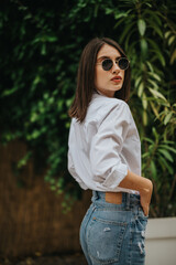 A stylish young woman outdoors, dressed casually in a white shirt and jeans, looking confident,...