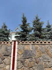 charming house partially hidden by tall fir trees, surrounded by sturdy stone fence. vibrant blue...