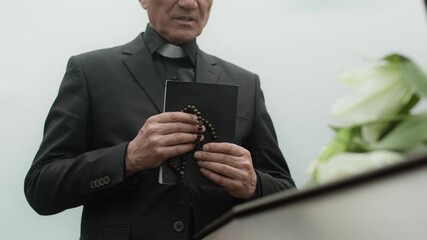 Midsection shot of mature priest holding Bible and rosary beads saying prayer standing in front of closed casket decorated with flowers during outdoor funeral ceremony