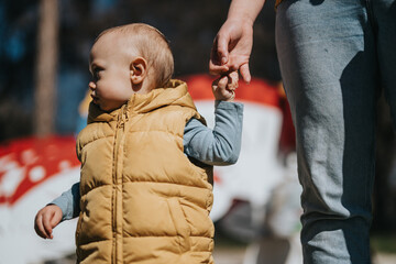 A young child in a yellow vest outdoors, holding an adult's hand and exploring during a warm, sunny...