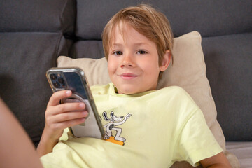 Portrait of a smiling blond child boy using smartphone sitting on the couch at home. Technology concept.