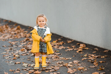 Smiling kid girl 3-4 year old wearing yellow raincoat and rubber boots walking in park with fallen...