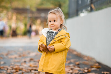 Smiling cute baby girl 3-4 year old wearing yellow raincoat walking in park with fallen leaves outdoors. Looking at camera. Autumn season. Childhood.